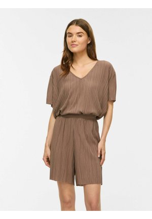 Femme aux longs cheveux bruns portant un haut marron plissé à col en V ample et un short plissé assorti sur un fond clair uni.
