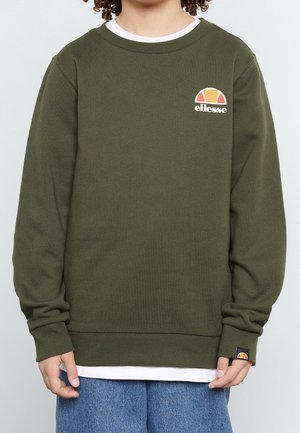 Person iført olivengrøn Ellesse sweatshirt over hvid skjorte og blå jeans, med krøllet hår delvist synligt.