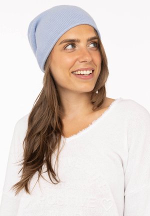 Light blue ribbed knit beanie worn with a white top featuring frayed neckline. Hair is long and wavy, with a neutral background.
