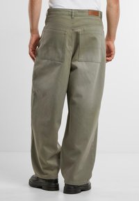 Baggy olive green canvas pants with two back pockets, visible stitching, and a brown leather label at the waistband. Pairing with black shoes.