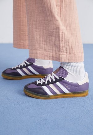 Persona que lleva zapatillas Adidas Gazelle moradas con rayas blancas, calcetines blancos y pantalones sueltos de color melocotón, de pie sobre un suelo azul.