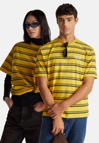 Žlutá trička s pruhy a černými linkami. Jedno tričko uvázané v pase. Oba modelové mají na sobě sluneční brýle a ležérní džíny.