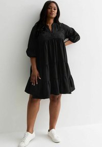 Vestido negro con diseño en capas, mangas cortas abullonadas y detalles bordados en el corpiño, combinado con zapatillas blancas sobre un fondo neutro.