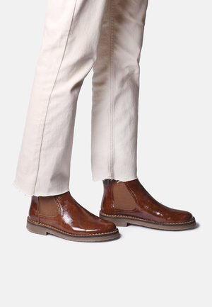 Bottines en cuir marron avec des panneaux élastiques sur les côtés, bout rond et coutures. Portées avec un pantalon blanc effiloché pour un contraste.