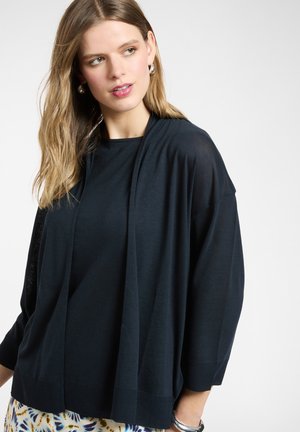 Joven con cabello largo castaño claro, lleva pendientes de aro plateados, un cárdigan negro suelto y pantalones estampados, mirando ligeramente hacia un lado.