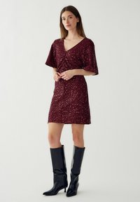 Burgundi spalvos blizgi suknelė su trumpomis rankovėmis, prigludusi silueto, mini ilgio, derinama su juodomis, iki kelių siekiančiomis, smailėmis botais.