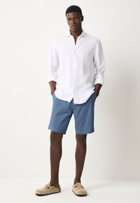 Bela lanena srajca z dolgimi rokavi, modri kratki hlači po meri in bež natikači. Preprost dizajn s sprednjimi gumbi in sproščenim krojem.