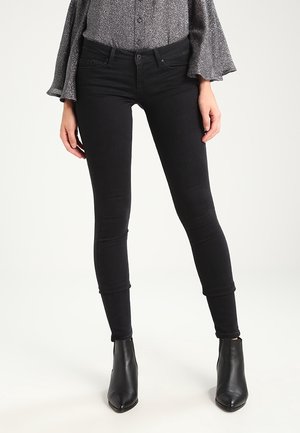 Mujer que lleva vaqueros negros ajustados combinados con botines negros y una blusa gris de manga larga con puños acampanados.