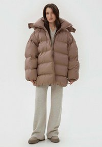 Brązowa pikowana kurtka o dużym, pudełkowatym kroju i wysokim kołnierzu. Posiada zamki błyskawiczne, duże puchy oraz prążkowane mankiety. Nosi się ją na szare spodnie dresowe i buty.