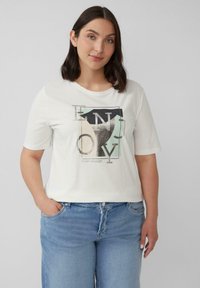 Frau mit schulterlangem, dunklem Haar, die ein weißes T-Shirt mit abstrakten Buchstaben und hellblauen Jeans trägt und mit einer Hand in der Tasche steht.