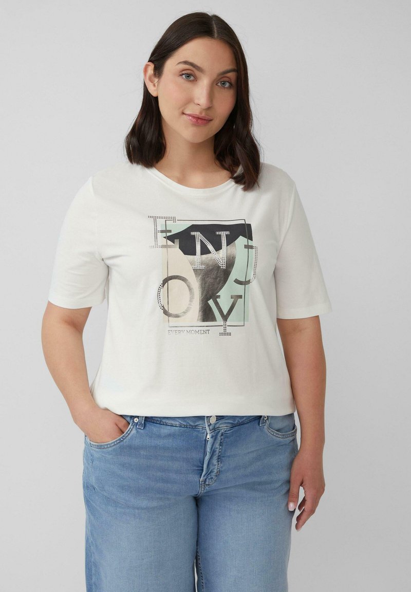Frau mit schulterlangem, dunklem Haar, die ein weißes T-Shirt mit abstrakten Buchstaben und hellblauen Jeans trägt und mit einer Hand in der Tasche steht.