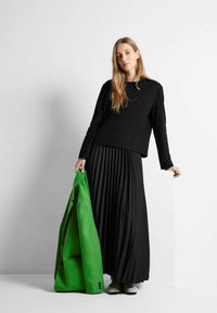 Schwarzes Langarmoberteil kombiniert mit einem schwarzen plissierten Maxir skirt. Hält eine lebhaft grüne Jacke. Die Schuhe sind metallic und haben ein rundes Zehendesign.