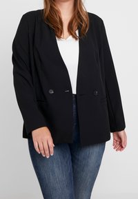 Mujer vestida con un blazer negro de doble botonadura sobre una blusa blanca y jeans azules, de pie contra un fondo claro y liso.