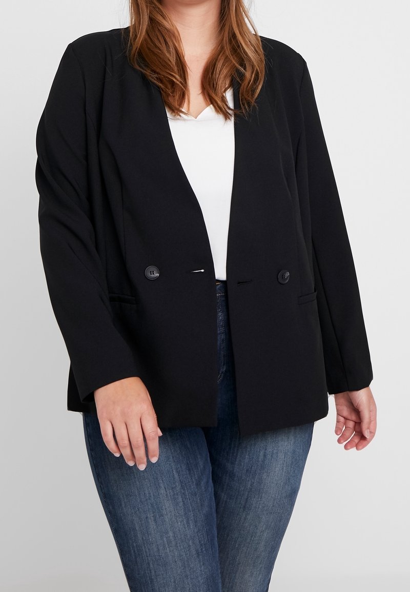 Mujer vestida con un blazer negro de doble botonadura sobre una blusa blanca y jeans azules, de pie contra un fondo claro y liso.