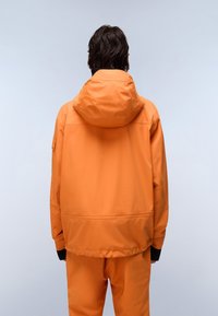 Personen iført en lys orange hættejakke og matchende bukser, set fra ryggen mod en ensfarvet lys baggrund.