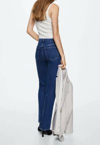 Jeans de tiro alto azules con una silueta ajustada, combinados con una blusa sin mangas blanca y una blazer gris claro colgada del brazo.