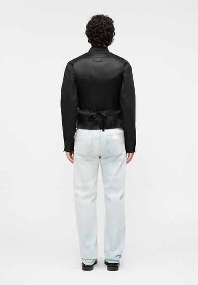 Μαύρο σατινέ σακάκι με σχέδιο δέσιμου στην πλάτη, μακριά μανίκια και δομημένο σχήμα, συνδυασμένο με ανοιχτόχρωμο τζιν και μαύρα παπούτσια.