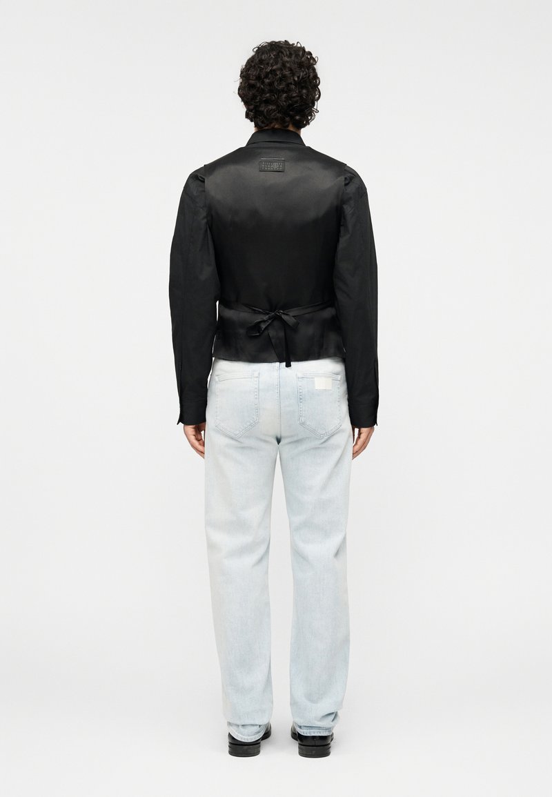 Veste en satin noir avec un design à nouer au dos, manches longues et forme structurée, associée à un jean bleu clair et des chaussures noires.