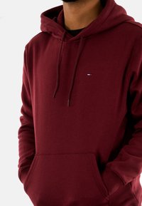 Bordeaux hoodie met een kangoeroezak, trekkoorden en een klein logodetail in blauw, wit en rood. Zachte stoftextuur.