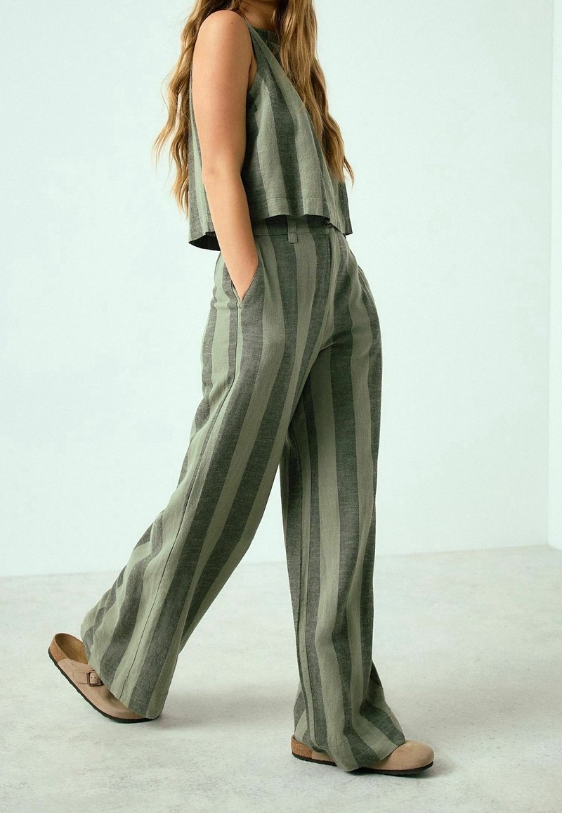 Femme portant un pantalon ample à rayures verticales vertes et grises et un haut sans manches assorti, debout les mains dans les poches.