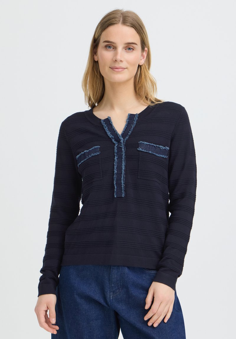 Femme aux cheveux blonds mi-longs portant un haut noir texturé à manches longues avec des finitions effilochées et un jean bleu, debout devant un fond clair.