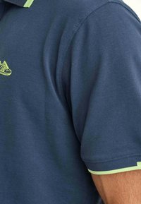 Mörkblå polotröja i texturerad tyg med limegrön krage och ärmaccenter; har en liten gul skologotyp på bröstet.