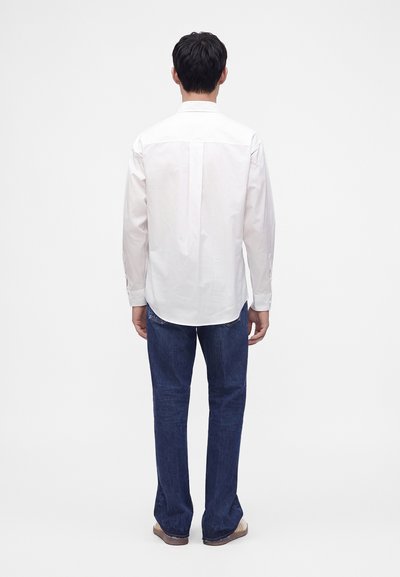 Homme de dos portant une chemise blanche à manches longues, un jean bleu et des chaussures marron sur un fond clair uni.