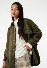 Olivgrüner, weicher Wildlederähnlicher Jacke mit Knopfverschluss, oversize Form und zwei Vordertaschen, kombiniert mit einem weißen Strickpullover und karierter Shorts.