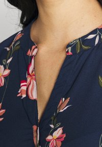 Marinebluse mit floralem Muster und Knopfleiste, die pinke und weiße Blumen sowie grüne Blätter zeigt. Weicher Stoff, mit Kragen und feinen Steppnähten.