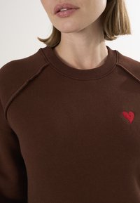 Bruine sweatshirt met een geribbelde ronde halslijn, subtiele schoudernaden en een kleine rode geborduurde hart op de voorkant. Zachte structuur.