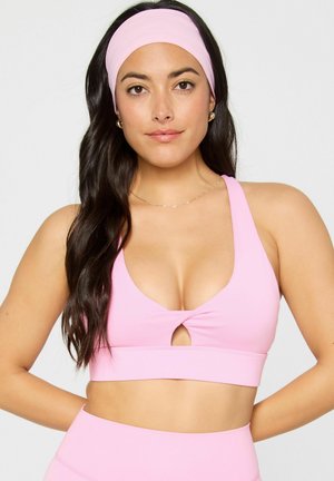 Woman with long dark hair wears a pink sports bra and matching headband, standing against a plain light background.