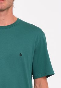 T-shirt en coton vert avec un col rond, des manches courtes et un petit emblème noir sur la poitrine. Texture douce et coupe décontractée.