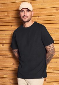 Černé tričko s kulatým výstřihem vyrobené z bavlny, s krátkými rukávy a volným střihem. Výrazné detaily zahrnují tetované ruce a světlou kšiltovku.