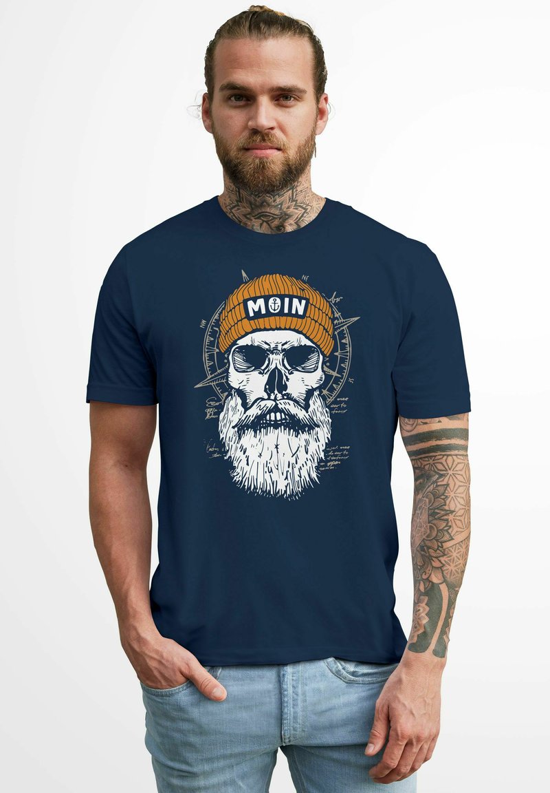 Navy blue t-shirt featuring a white graphic of a skull with a beard and a tan knit beanie that reads "MOIN." Smooth cotton fabric.