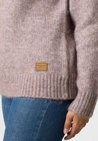 Lichtroze gebreide trui met een gestructureerde afwerking, geribbelde manchetten en zoom, met een bruin leren label met "SWEDEMOUNT." Gestyled met blauwe jeans.