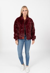 Giacca bomber in pelliccia sintetica bordeaux con polsini e orlo a coste, zip frontale e tasche laterali; indossata su un top nero e abbinata a jeans.
