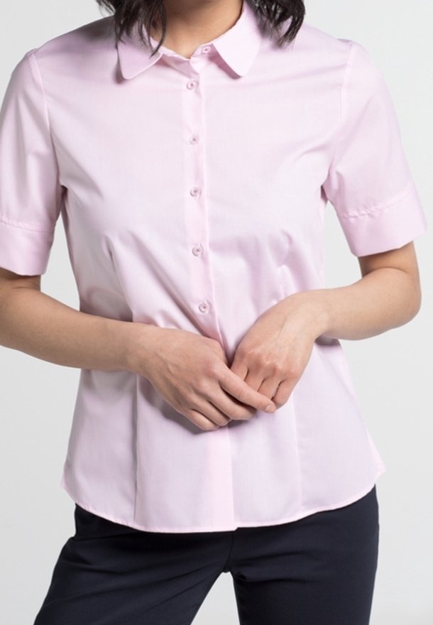 Femme portant une chemise à manches courtes rose clair avec col et boutons, mains doucement jointes devant, assortie à un pantalon foncé.