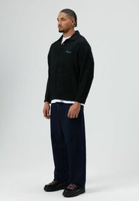 Schwarzer, strukturierter Fleecepullover mit Kragen, kombiniert mit weiten dunkelblauen Jeans und schwarzen Schuhen mit pinken Schnürsenkeln und einer klobigen Sohle.