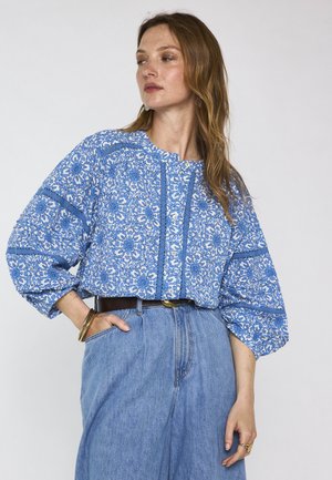 Vrouw met blauwe blouse met patroon en ballonmouwen, lichtblauwe hoge taille jeans, zwarte riem en gouden armbanden, staand met hand in zak.