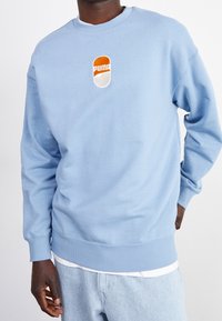 Sudadera azul claro hecha de una mezcla de algodón. Presenta un logotipo de Puma bordado en naranja y blanco en el pecho. Puños y dobladillo acanalados.
