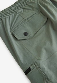 Shorts vert olive avec ceinture élastique, dotés d'une poche à rabat avec fermeture à bouton-pression noir et d'une poche zippée sur le côté. Texture lisse.