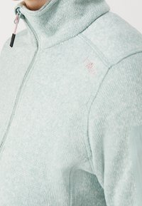 Gros plan d'une personne portant un pull zippé texturé bleu clair avec un col montant et un curseur de fermeture éclair rose.