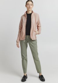Lichtroze fleecedwz jacket met ritssluiting en een staande kraag, twee zakken, gecombineerd met olijfgroene broek en zwarte instappers. Glad textuur, casual ontwerp.