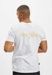Hvid bomulds t-shirt med ordet "mentalité" i guld og "CAYLER" i blå på ryggen. Korte ærmer og almindelig pasform.