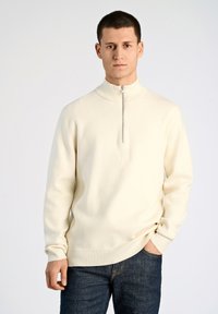 Krämfärgad halvziptröja i mjukt tyg med en åtsittande krage och ribbade muddarna, tillsammans med mörka denimjeans.