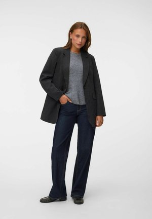 Femme debout portant un blazer gris charbon, un pull gris, un jean bleu foncé, des chaussures noires et des boucles d'oreilles en perles sur un fond blanc uni.
