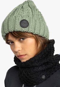 Mujer con un gorro de punto verde claro y un grueso bufanda de punto negra sobre una chaqueta oscura, mirando ligeramente hacia la izquierda.