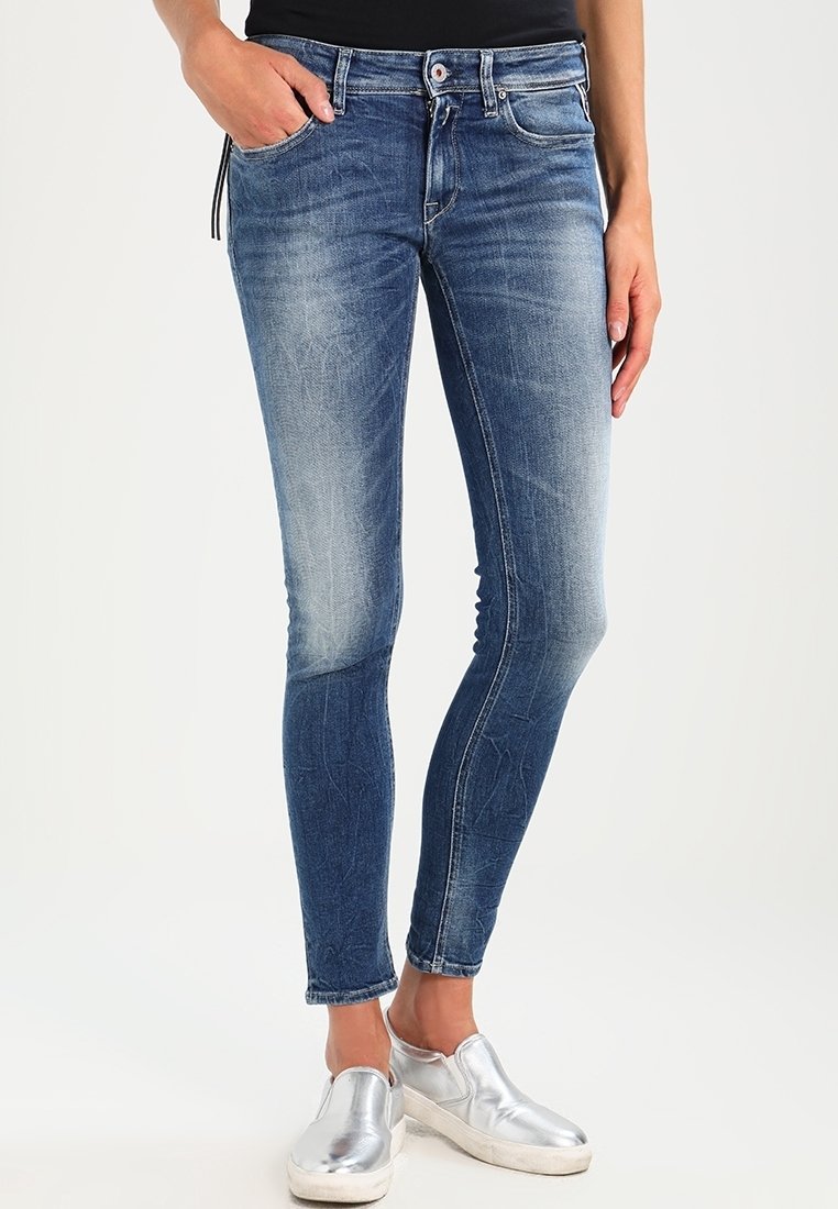 Mujer vestida con jeans ajustados azul desgastados, con una mano en el bolsillo y zapatos metálicos plateados tipo slip-on, de pie frente a un fondo sencillo.
