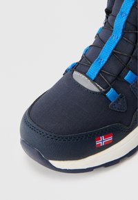 Sötétkék sportcipő texturált anyagból, kék és fekete fűzőkkel, gumitalppal, és egy kis norvég zászlóval a oldalon.