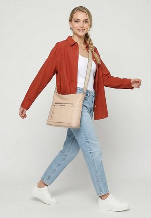 Joven camina con una camisa roja abierta, camiseta blanca sin mangas, jeans azul claro, zapatillas blancas y bolso cruzado beige con bolsillo con cremallera.